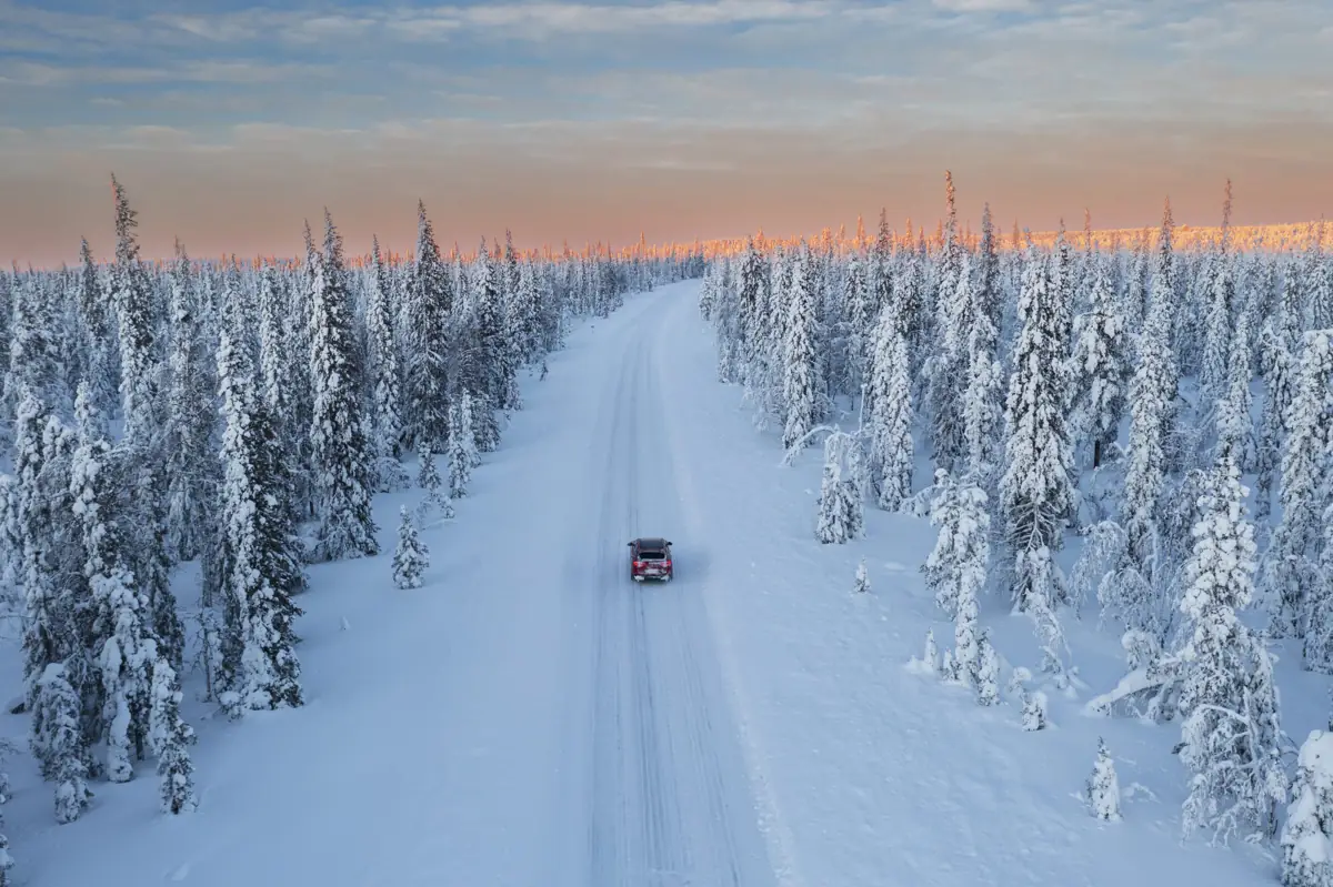 An aerial view of a lone car on a snowy road through the frozen forests of Lapland at soft winter sunset